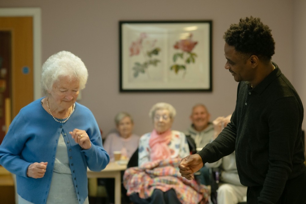 An elderly woman and a man in a black shirt dancing, with three other elderly people seated in the background.
