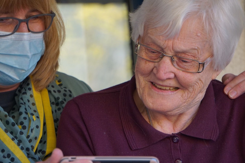 A smiling elderly woman in glasses looks at a smartphone held by a woman in a mask and glasses.