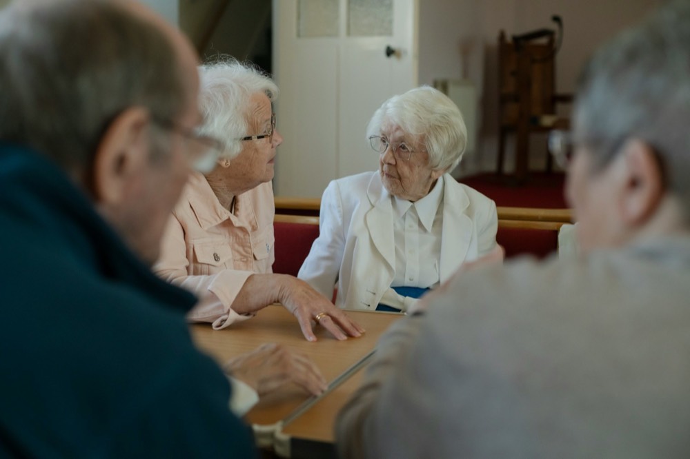 Four elderly people gathered around a table, two women facing each other, two blurred in the foreground.