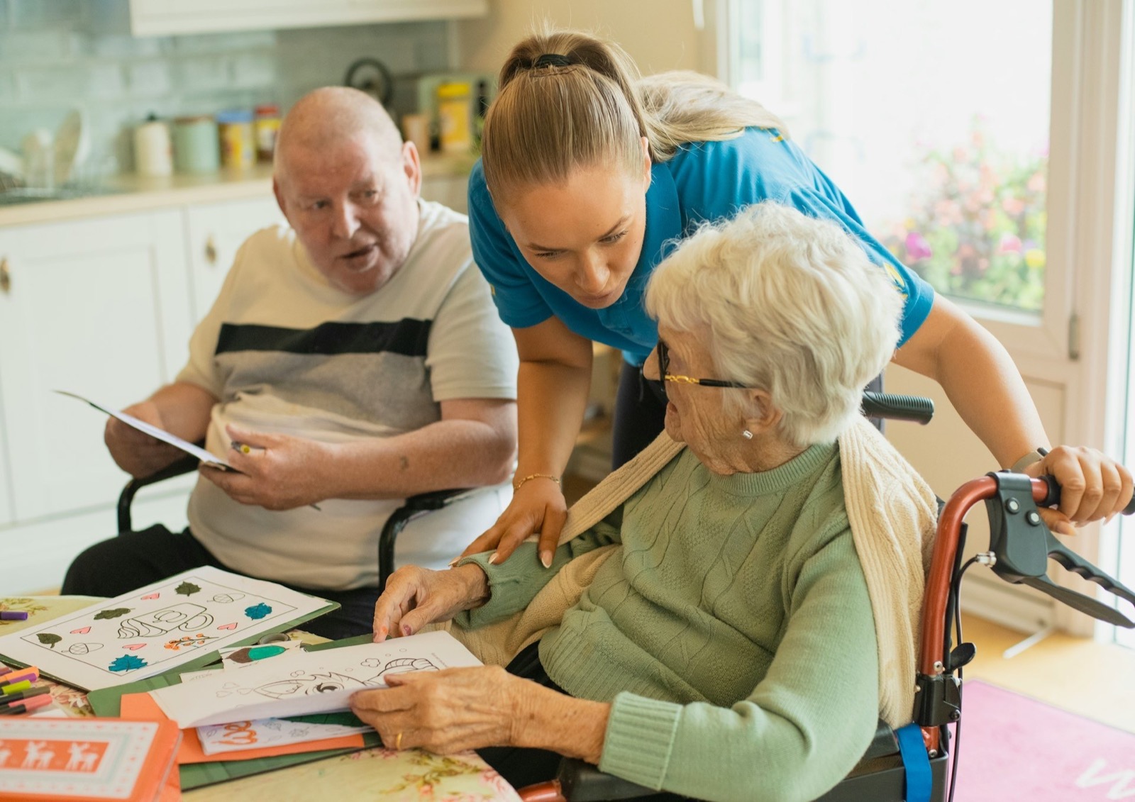 Caregiver and two seniors coloring at a table; caregiver points to a drawing, helping an elderly woman.