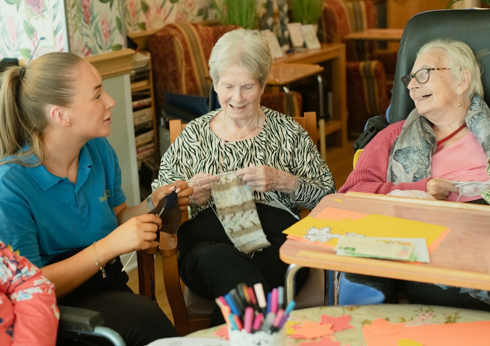 Three women, two elderly and one younger, sit around a table, smiling and engaged in a craft activity.