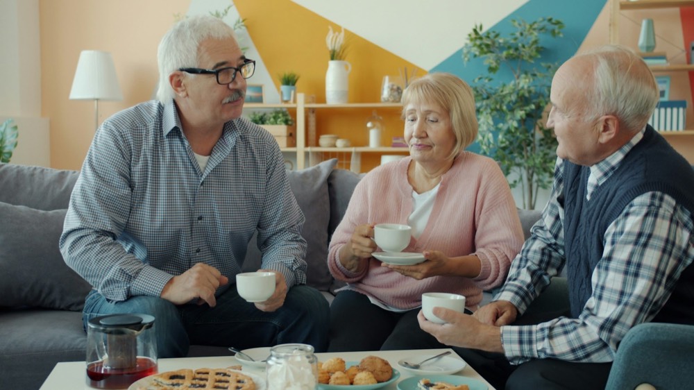 Three seniors, two men and one woman, sit on a sofa, drinking tea and conversing in a living room.